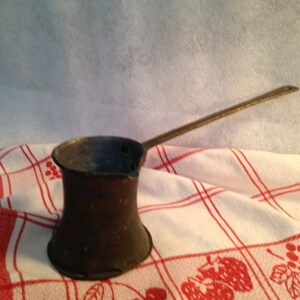 May include: A dark, weathered metal coffee pot with a wide base, a narrow neck, and a long, slender handle extending from the side. The pot features a small pouring spout at the rim and shows signs of oxidation and age on its exterior surface.
