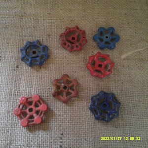 Könnte beinhalten: Sieben antike Wasserhahn-Griffe aus Metall in verschiedenen Blautönen und Rot. Die Griffe sind sternförmig mit einem zentralen Loch und auf einem Jutesack-Hintergrund angeordnet.