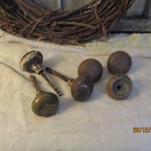 Może przedstawiać: Sześć antycznych gałek drzwiowych z różnymi wykończeniami, w tym mosiądz, brąz i biała farba. Gałki znajdują się na białej powierzchni tkaniny.