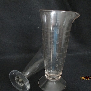 Puede incluir: Dos cilindros graduados de vidrio transparente con una base ancha. El cilindro de la derecha tiene marcas hasta 8 onzas. El cilindro de la izquierda es más pequeño y de forma cónica.