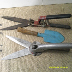 May include: Three gardening tools: a pair of red and black pruning shears, a blue and silver pair of shears with the text "Wizard Garden Shears", and a small blue trowel with a wooden handle.