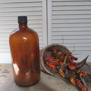 May include: A large, amber-coloured glass bottle with a black lid. The bottle is sitting on a hessian surface next to a cornucopia filled with autumn foliage and berries.