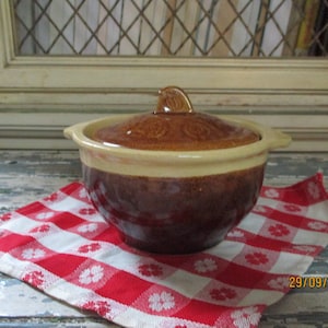 以下が含まれることがあります： 茶色とベージュのセラミック製のキャセロール鍋と蓋。鍋は茶色の釉薬で、ベージュの縁取りが施されています。蓋には鳥の形をしたつまみがあります。