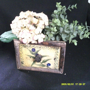 May include: A vintage wooden box with a painted floral design featuring blue berries and green leaves. The box is decorated with a yellow background and a brown wooden frame. The box is filled with white hydrangea flowers and green eucalyptus.