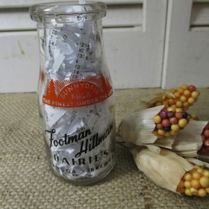 Puede incluir: Una botella de leche de vidrio transparente con una etiqueta de papel blanco que dice "Sunnydale Milk The Finest Under The Sun Footman Hillman Dairies Bangor, Brewer". La botella está llena de trozos de papel blanco. La botella está sentada sobre una superficie de arpillera marrón junto a un montón de mazorcas de maíz artificiales.