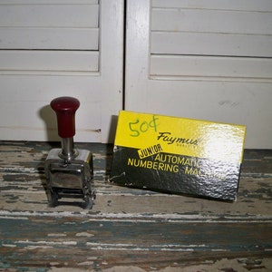Puede incluir: Un sello numerador vintage de color plateado con un mango rojo. El sello está etiquetado como "Famous" y está en una caja amarilla que dice "Junior Automatic Numbering Machine".