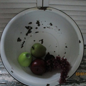 May include: A white enamel bowl with brown rust spots and a black rim. The bowl contains three green apples, a red apple, and a bunch of red grapes.
