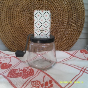 May include: A vintage glass and metal hand-crank food grinder with a black base and a white and black patterned lid. The grinder is sitting on a white and red checkered tablecloth.