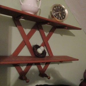 May include: A wooden wall shelf with a criss-cross design. The shelf has two levels and is painted a reddish brown colour. There is a small bird's nest with two white eggs on the lower shelf. A white ceramic jug and a gold clock are on the top shelf.