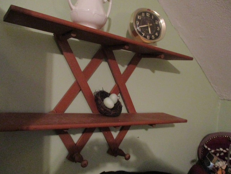 May include: A wooden wall shelf with a criss-cross design. The shelf has two levels and is painted a reddish brown colour. There is a small bird's nest with two white eggs on the lower shelf. A white ceramic jug and a gold clock are on the top shelf.