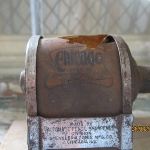 May include: Vintage metal pencil sharpener with a rusty brown finish. The sharpener has a label that reads "Chicago Made by Spengler Loomis Mfg. Co. Chicago, Ill."