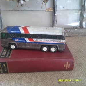 Peut inclure: Un bus jouet Greyhound vintage peint en rouge, blanc et bleu. Le bus est posé sur un livre rouge avec le titre "Radiant Heating Adlam" visible.