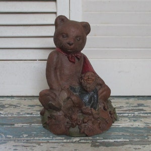 May include: A brown ceramic bear figurine with a red bow tie sitting with a small gnome figurine. The bear and gnome are sitting on a base of brown ceramic with leaves and flowers.
