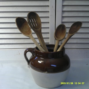 May include: A brown and white ceramic crock with a handle, filled with wooden spoons. The spoons are various sizes and shapes, including a slotted spoon.