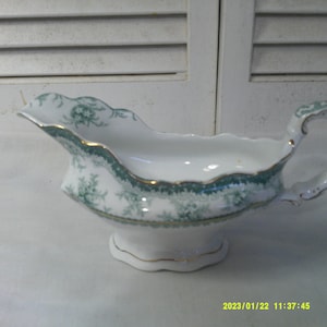 Op de afbeelding: Een witte porseleinen sauskom met een groen en gouden bloemenmotief. De kom heeft een gegolfde rand en een handvat.