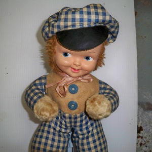 May include: A vintage plush toy boy doll wearing a blue and white checkered hat and matching pants. The doll has a smiling face with blue eyes and a brown fur trim.