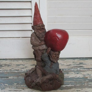 Könnte beinhalten: Zwei Gnome, einer mit roter Mütze, halten einen großen roten Apfel. Die Gnome sind aus braunem, strukturiertem Material gefertigt und stehen auf einem braunen Sockel.