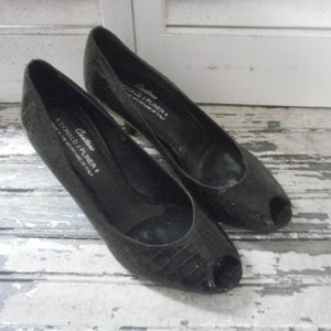 May include: A pair of black peep-toe heels. The shoes have a textured, reptile-like pattern and a low heel. The brand name "DONALD J PLINER" is visible on the side of one shoe. The shoes are displayed on a distressed, white-painted wooden surface.