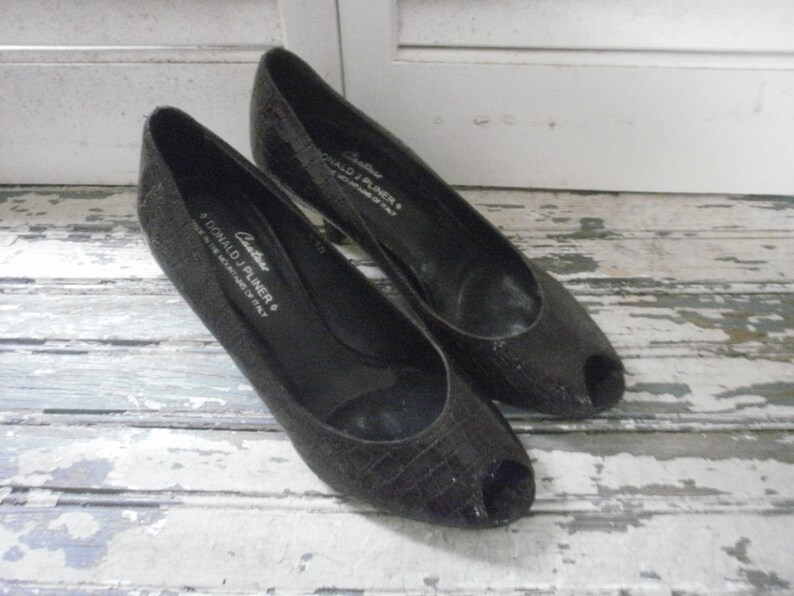 May include: A pair of black peep-toe heels. The shoes have a textured, reptile-like pattern and a low heel. The brand name "DONALD J PLINER" is visible on the side of one shoe. The shoes are displayed on a distressed, white-painted wooden surface.