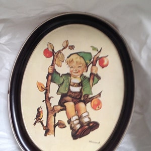 May include: Oval framed print featuring a smiling child in traditional Bavarian clothing, perched on a tree branch with apples. The frame is dark brown with a silver inner edge. A small bird is perched on the tree.