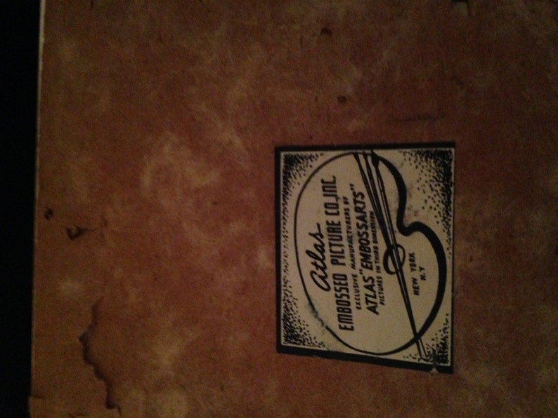 May include: A vintage label from Atlas Embossed Picture Co. Inc. is affixed to a textured, brown surface. The label features text and a stylized artist's palette, with the words "Atlas Embossed Picture Co. Inc." and "New York, N.Y."