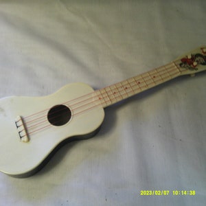 Puede incluir: Un ukelele blanco con cuerdas rojas y un diseño de personaje de dibujos animados en la pala.