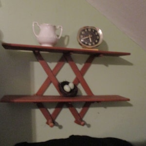 May include: A wooden shelf unit with a criss-cross design. The shelf has two tiers and is mounted on a wall. A white jug, a clock, and a bird's nest with two eggs are on the shelves.