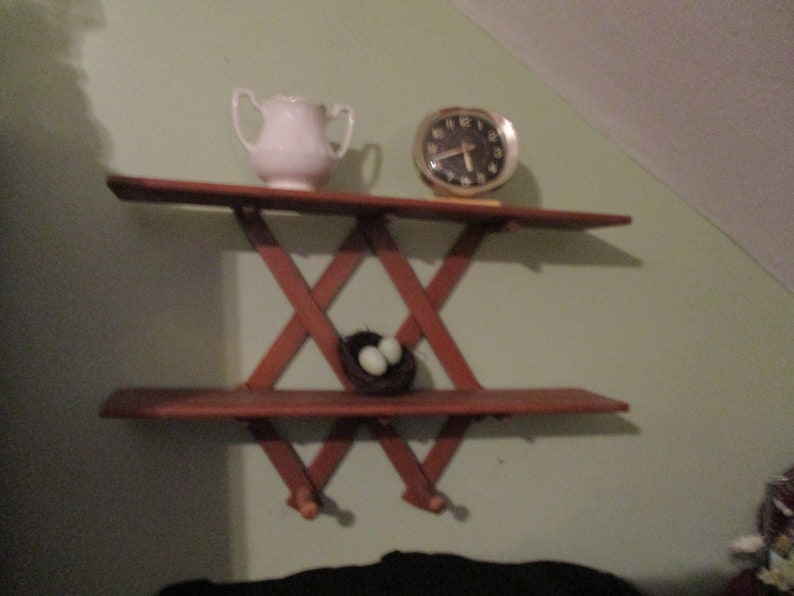 May include: A wooden shelf unit with a criss-cross design. The shelf has two tiers and is mounted on a wall. A white jug, a clock, and a bird's nest with two eggs are on the shelves.