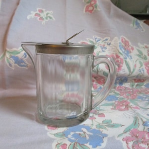 May include: A clear glass pitcher with a silver lid and a handle. The pitcher is sitting on a floral tablecloth.