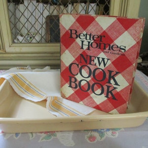 May include: A vintage Better Homes and Gardens New Cookbook with a red and white gingham cover. The cookbook is sitting on a light beige baking dish.