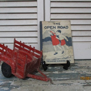 May include: A red toy wagon with a wooden bed and black wheels sits next to a vintage children's book titled "The Open Road" with a colorful illustration of two children walking on a path.