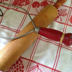 Puede incluir: Un rodillo de madera con mango rojo y un reposa-batidor de metal sobre un mantel a cuadros rojo y blanco. El rodillo es de color marrón claro y el reposa-batidor es plateado.
