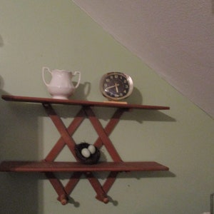 May include: A wooden shelf with a criss-cross design, featuring a white ceramic jug, a gold clock with black hands, and a bird's nest with two blue eggs.