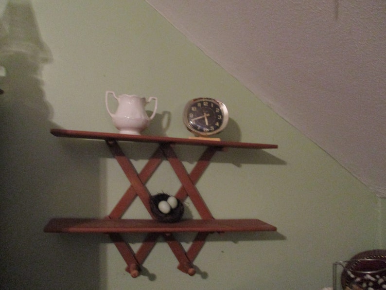 May include: A wooden shelf with a criss-cross design, featuring a white ceramic jug, a gold clock with black hands, and a bird's nest with two blue eggs.