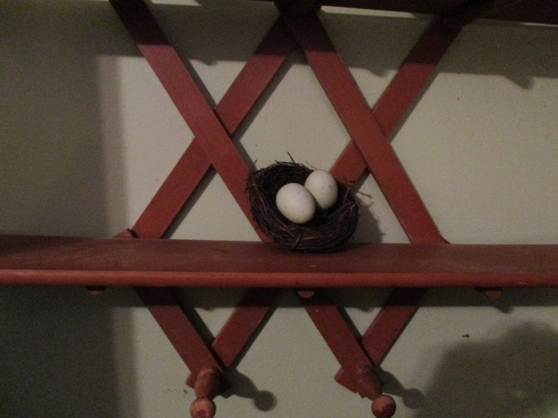May include: A wooden shelf with a criss-cross design, painted red. A small bird's nest with two white eggs sits on the shelf.
