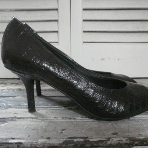 May include: A pair of black high-heeled shoes with a textured, reptile-like pattern. The shoes have pointed toes and slender heels. The heels are approximately 3 inches tall.