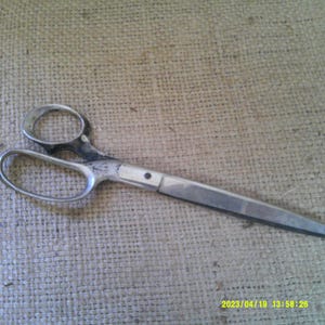 May include: Vintage metal scissors with silver-colored handles and blades. The scissors have a classic design with large finger holes and a pointed tip. The blades show signs of wear, suggesting they have been used.