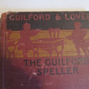 May include: A vintage red book with gold lettering titled "The Guilford Speller" by Guilford & Lovell. The cover features a black and gold illustration of two figures carrying a wheelbarrow.
