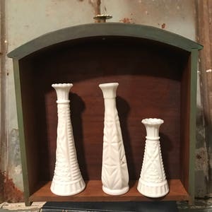 May include: Three white milk glass bud vases with a geometric pattern, displayed in a green painted wooden shelf with a brass knob. The shelf is sitting on top of two black books with gold lettering.