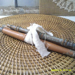 May include: Two antique wooden spindles with metal accents, wrapped with a piece of white fabric. The spindles are brown and copper-colored, resting on a woven wicker surface. The background includes a window and a burlap-like material.