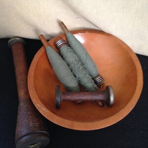 May include: A wooden bowl filled with three spools of green thread and a wooden spindle. The bowl is sitting on a beige fabric surface.