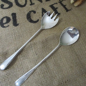 May include: A set of two silver-plated serving utensils, a fork and a spoon, with long handles. The utensils are lying on a hessian surface.