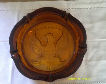 May include: Amber glass ashtray with an embossed design of an American bald eagle with wings spread, holding a banner with stars and stripes. The eagle is perched on a branch with leaves.