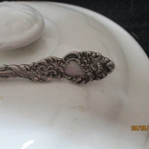 Puede incluir: Una cuchara de plata con un diseño ornamentado. La cuchara tiene un cuenco en forma de corazón y un mango retorcido.