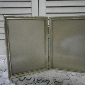 May include: A silver-toned metal picture frame with two hinged openings. The frame has a decorative border and is designed to hold two photos.