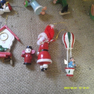 May include: A set of vintage wooden Christmas decorations. The decorations include a clock, a Father Christmas, a hot air balloon, and a small figure. The decorations are painted in red, white, green, and blue.