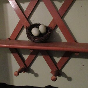 May include: A wooden shelf with a criss-cross design, painted red, is mounted on a wall. Two light blue eggs are nestled in a brown bird's nest on the shelf.