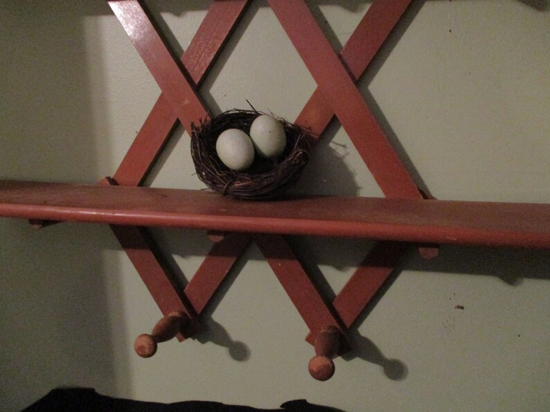 May include: A wooden shelf with a criss-cross design, painted red, is mounted on a wall. Two light blue eggs are nestled in a brown bird's nest on the shelf.