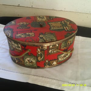 May include: A vintage oval-shaped hat box with a red base and a colourful patterned lid. The lid features a variety of images, including people, animals, and buildings. The box has a rope handle.