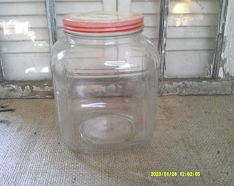 May include: A clear glass jar with a red and white lid. The jar has a square shape with rounded corners and a textured pattern on the sides. The jar is sitting on a burlap surface.